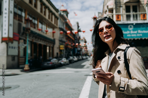 Happy young woman in sunglasses enjoying weekends spending time outdoors holding smartphone for chatting. cheerful girl laughing on street looking away mobile phone in hand walking in chinatown. © PR Image Factory