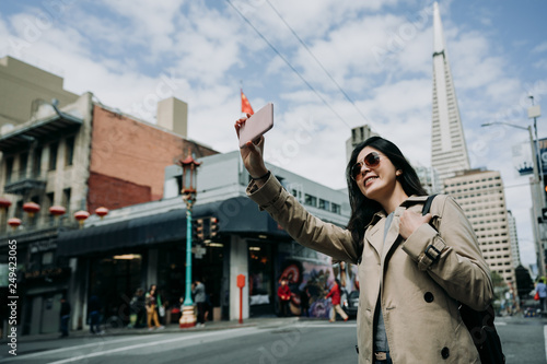young girl taking selfie standing on road in chinatown. female asian travel backpacker holding cellphone make self portrait with Transamerica Pyramid tower building in san francisco in usa.