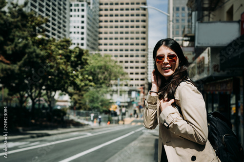 young asian lady backpacker in sunglasses face camera smiling charming. girl tourist laughing carrying bag standing on street with trees in park in spring. happy woman on road in busy shopping area.