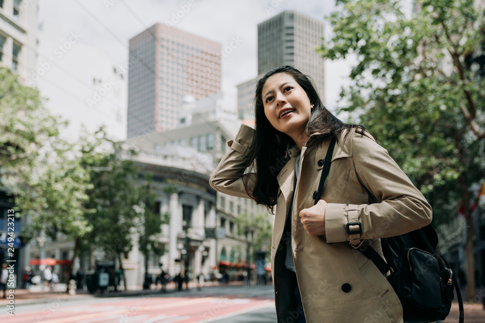 Fototapeta premium asian traveler woman smiling cheerful standing on street with green trees on sidewalk in spring. young girl local lifestyle go to work in morning san francisco usa. female backpacker sightseeing city