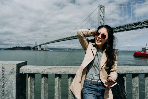 travel asian woman charming smiling relying on handrail holding smart phone. boat sail on water blue clear ocean in background under oakland bay bridge san francisco on sunny day wearing sunglasses.