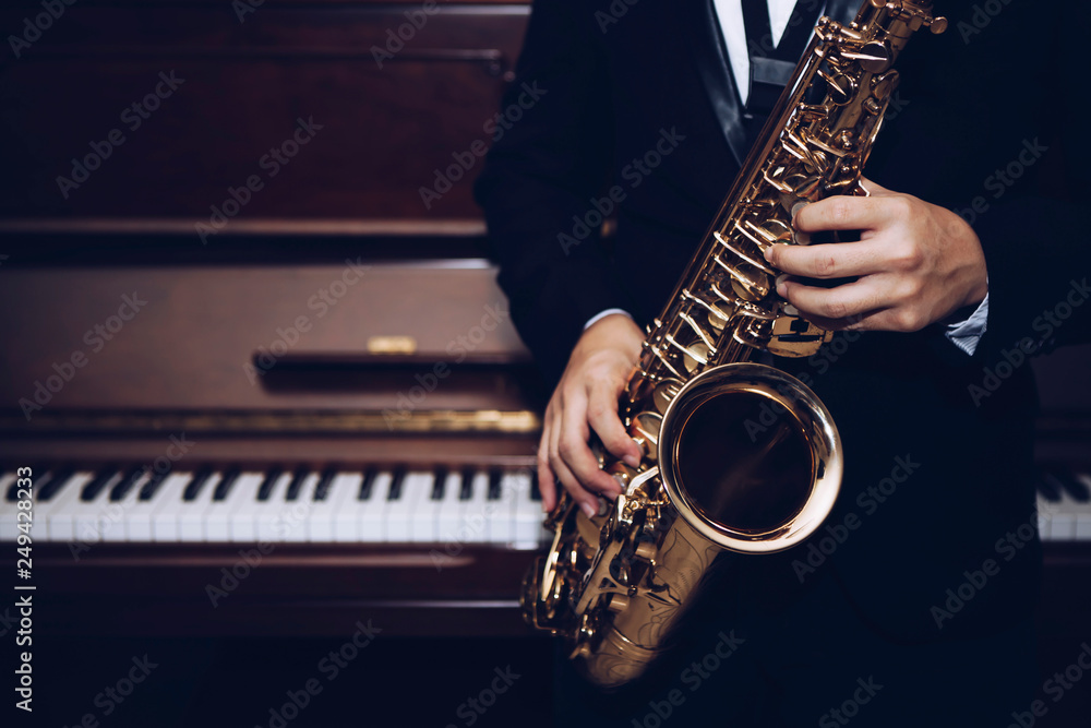Obraz premium close up of Young Saxophone Player hands holding alto sax musical instrument with piano background in dark room, vintage tone, can be used for music background, copy space