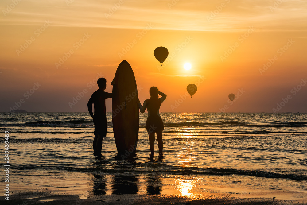Silhouette of Surfer man and younger sister standing happily and enjoy on the beach looking out to hot air balloon over the sea with surfboards in the sunset after surfing.
