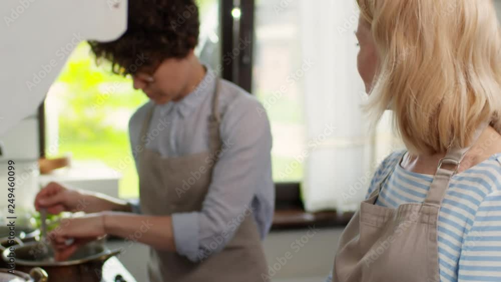 Tilt up of middle-aged blonde woman making buns from dough and talking to young woman with short curly hair mixing soup with spoon and salting it Tilt up of middle-aged blonde woman making buns from dough and talking to young woman with short curly hair mixing soup with spoon and salting it