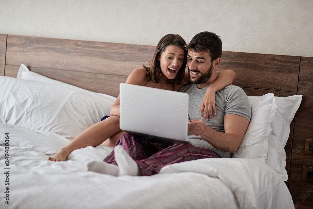 Happy couple lying on a bed with computer - Beautiful married couple watching sex video on laptop laughing together - People, sexual, technology concept.