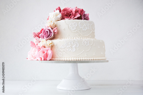 Two-tiered white wedding cake decorated with pink flowers on a white wooden background.