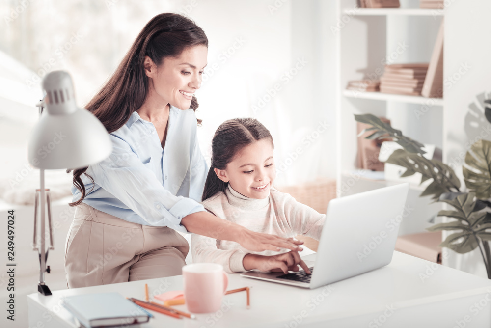 Caring smiling mother pointing at laptop showing daughter their family photos Caring smiling mother pointing at laptop showing daughter their family photos