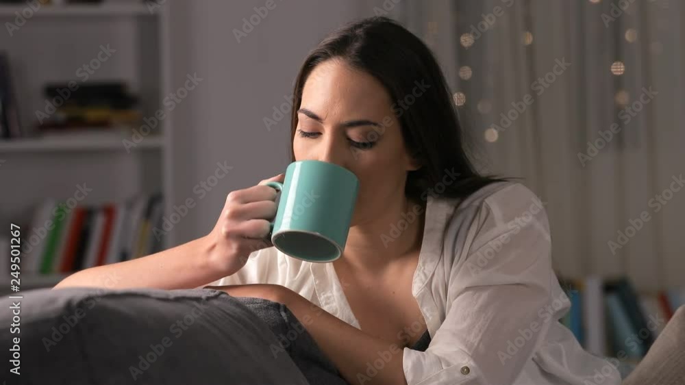 Serious pensive woman drinking coffee sitting on a couch at home in the night