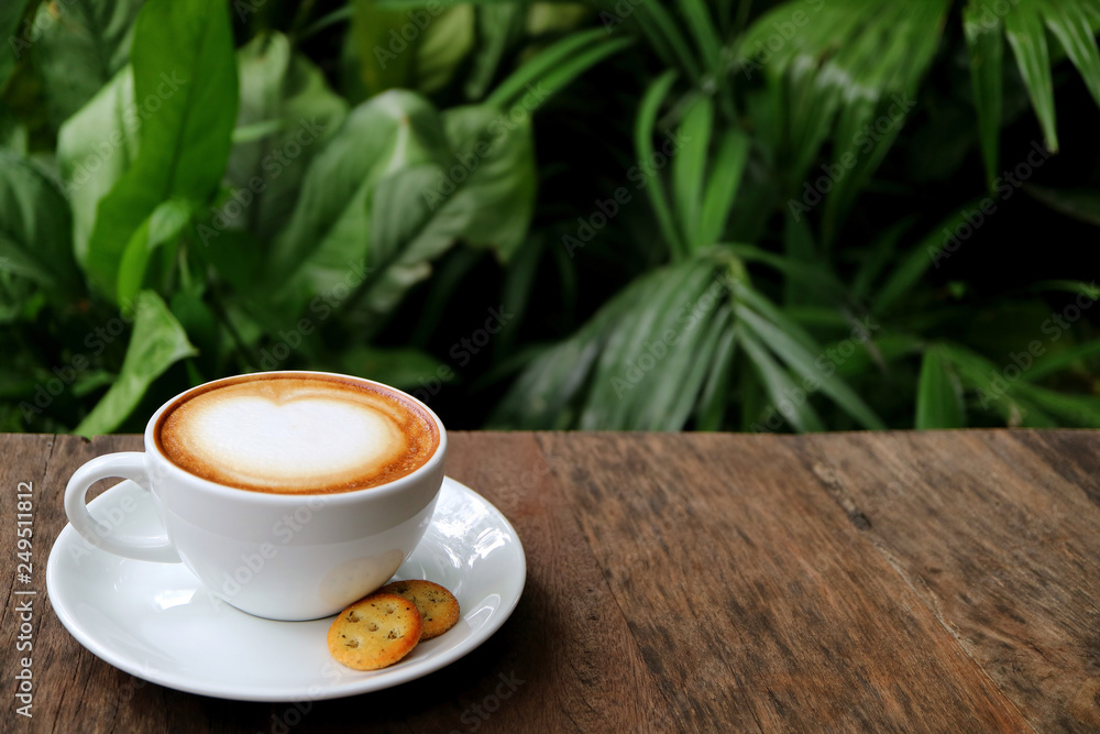 Obraz premium Cup of Hot Cappuccino Coffee with Cookies Served on Wooden Table with Blurry Green Foliage in Background