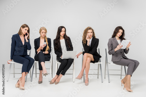 attractive colleagues in formal wear sitting on chairs and using gadgets isolated on grey