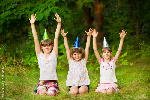 Three friendly children in festive cone caps, sit on grass, have fun together as celebrate birthday look with happy expressions at camera