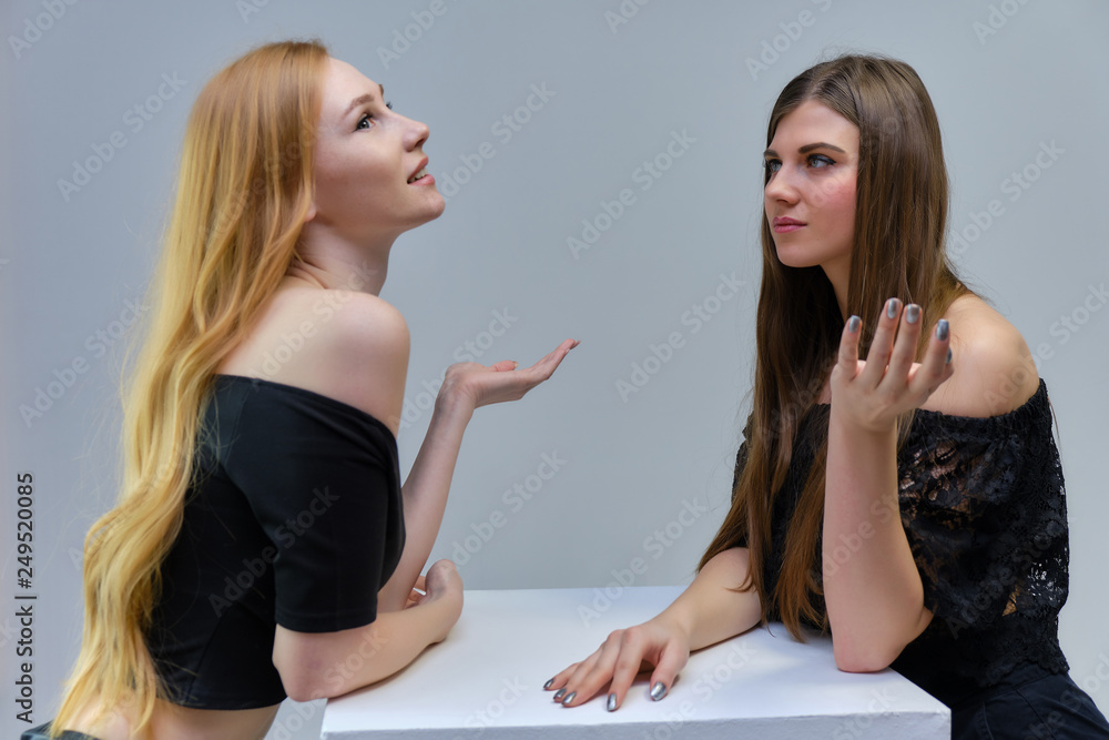 Concept portrait of two cute beautiful girls talking on a gray background in the studio standing at the table Concept portrait of two cute beautiful girls talking on a gray background in the studio standing at the table