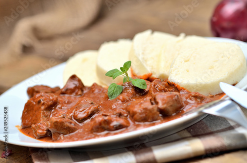 Close-up of pork goulash meat with dumplings on white plate, cutlery, garlic, onion, pepper, tablecloth in the background - typical Czech food