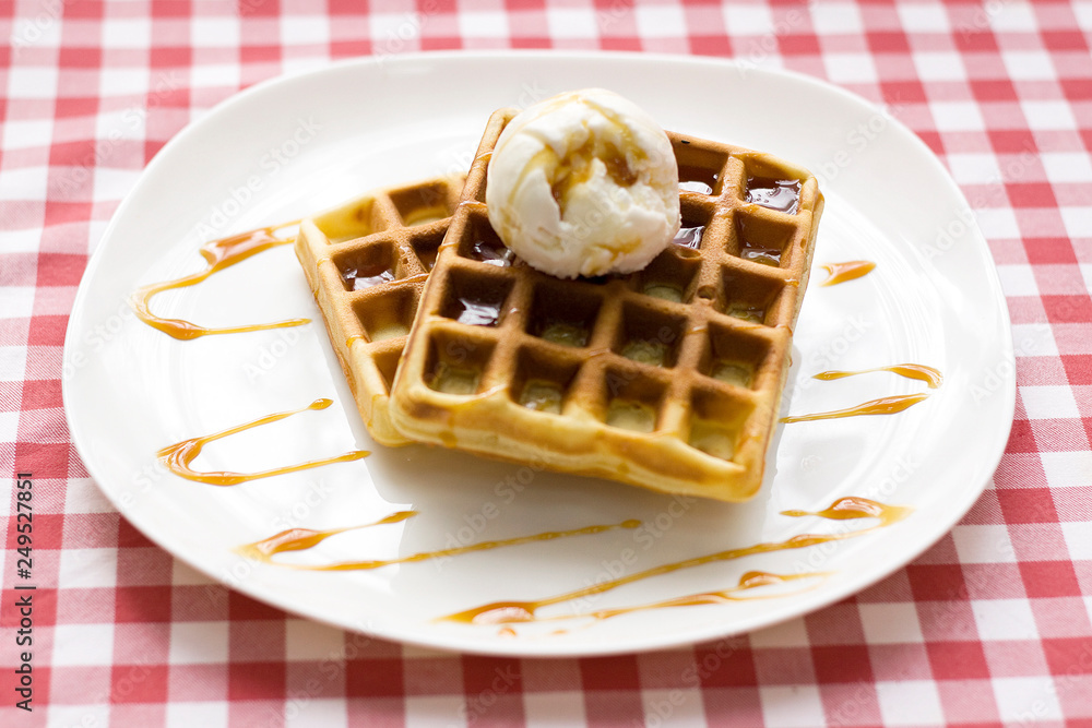 Plate of belgian waffles with ice cream and caramel sauce on red checkered tablecloth