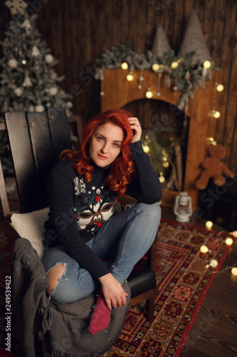 Attractive happy young woman in cozy comfortable clothes is sitting near a Christmas tree