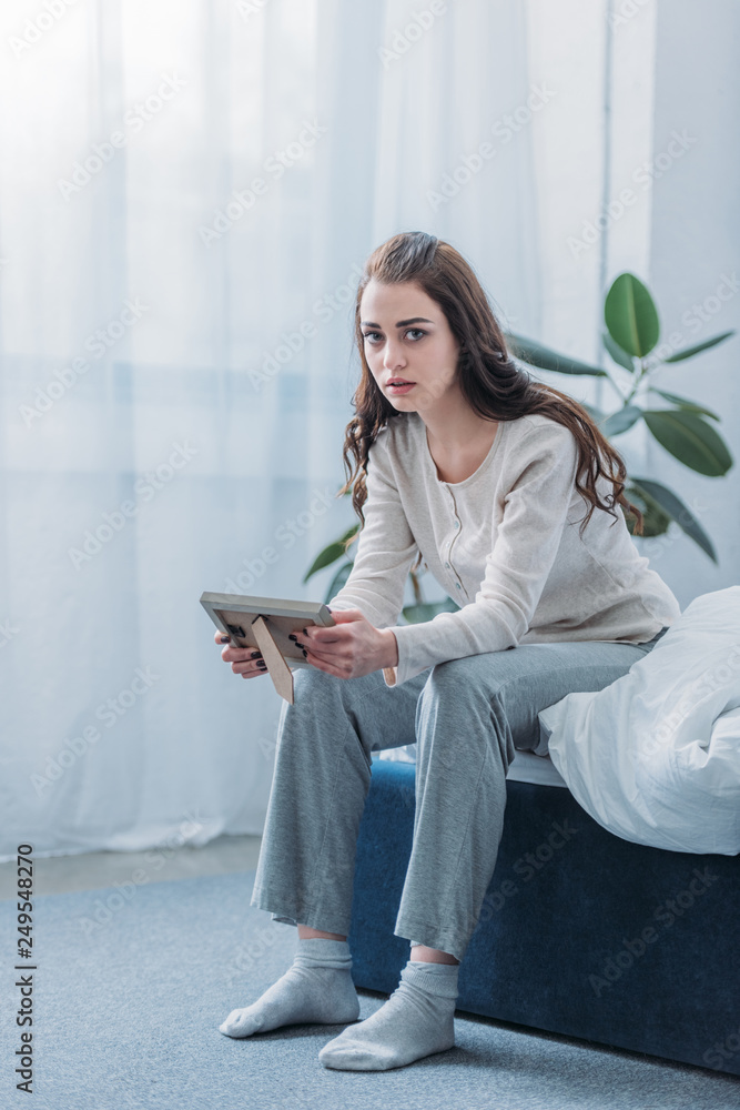grieving woman sitting on bed, looking at camera and holding picture frame at home grieving woman sitting on bed, looking at camera and holding picture frame at home