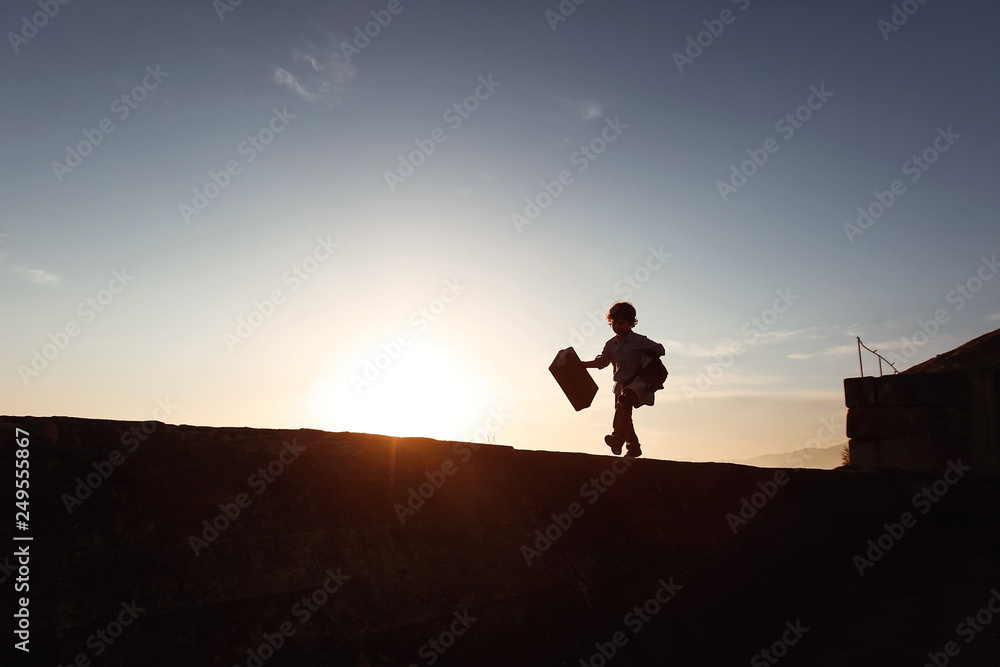 A little boy runs along a ledge at sunset with a suitcase in his hand. Little traveler. Tourism. Vintage. Escape from home. Silhouette.