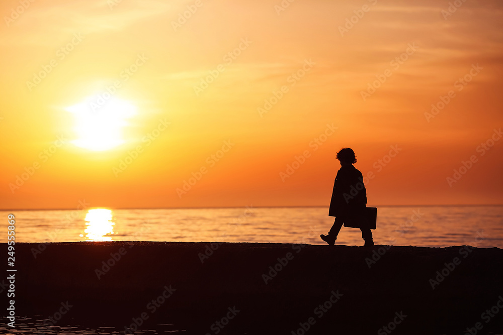 A little boy in a coat with a suitcase, walking along the pier at sunset. Little traveler. Tourism. Vintage. Escape from home. Silhouette.