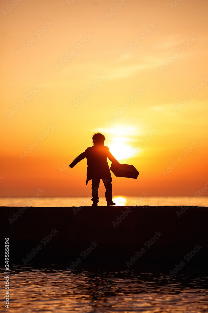 A little boy in a coat with a suitcase, walking along the pier at sunset. Little traveler. Tourism. Vintage. Escape from home. Silhouette.
