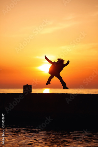 A little boy in a coat with a suitcase  jumping with happiness on the pier at sunset. Little traveler. Tourism. Vintage. Escape from home. Silhouette.