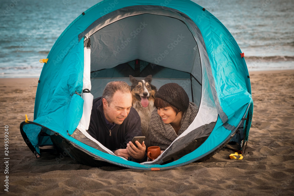 couple looking at the smart phone and have fun inside a tent in free camping on the beach Dog border collie behind them looking at the camera. bright colors and alternative vacation family concept couple looking at the smart phone and have fun inside a tent in free camping on the beach Dog border collie behind them looking at the camera. bright colors and alternative vacation family concept