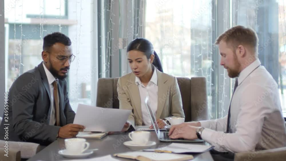 Young Asian businesswoman sitting at table in cafe with two male colleagues and discussing document with middle eastern man while Caucasian businessman working on laptop Young Asian businesswoman sitting at table in cafe with two male colleagues and discussing document with middle eastern man while Caucasian businessman working on laptop