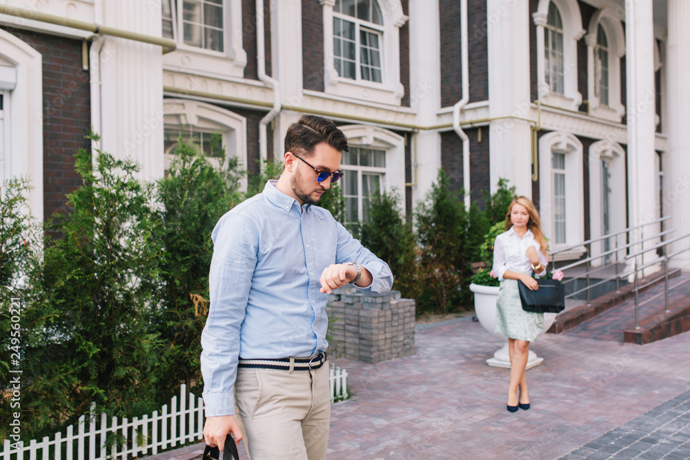 Handsome businessman in sunglasses looking at watch on street. Pretty elegant blonde girl is waiting for him on background Handsome businessman in sunglasses looking at watch on street. Pretty elegant blonde girl is waiting for him on background