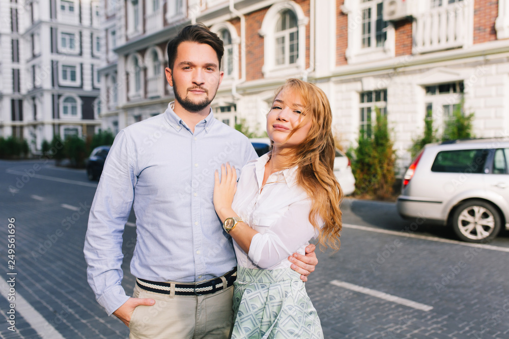 Portrait of cute couple walking around British quarter. Handsome guy looking seriously, blonde girl smiling to camera