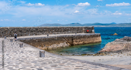 One man standing on a large stone pier on a sunny day, with blue ocean and mountains in the background. Taken in Renvyle, along the Wild Atlantic Way in Ireland in summer.