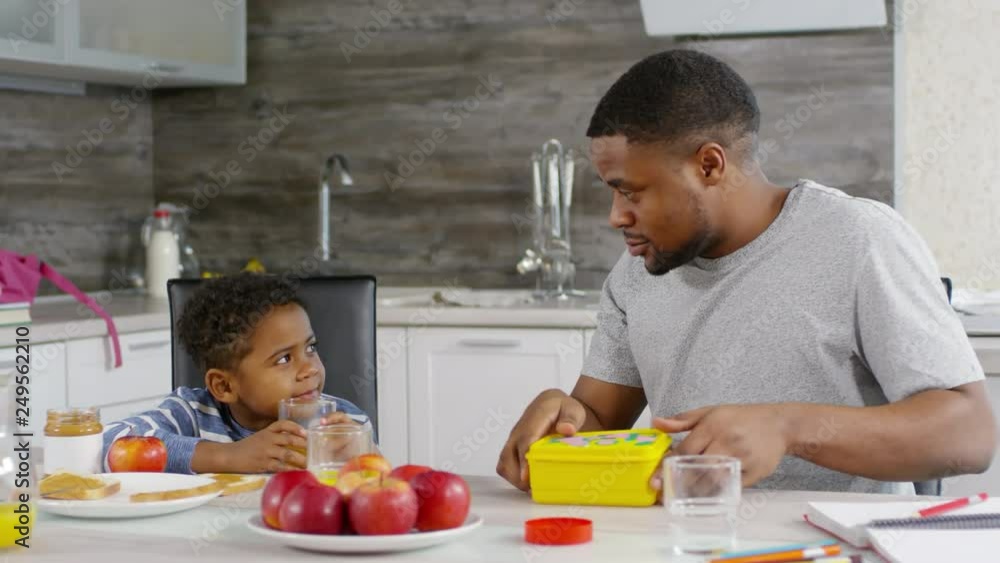 Lockdown of African man preparing school snack box for his son in home kitchen while talking to him, then helping him putting on school backpack Lockdown of African man preparing school snack box for his son in home kitchen while talking to him, then helping him putting on school backpack