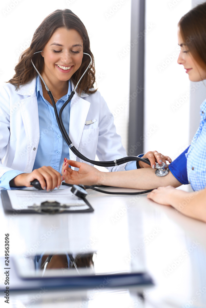 Doctor checking blood pressure of female patient while sitting at the desk in hospital office. Cardiology in medicine and health care concept Doctor checking blood pressure of female patient while sitting at the desk in hospital office. Cardiology in medicine and health care concept