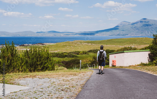 Man with a backpack, cap & shorts walking down a hill, with mountains & ocean in the distance. Taken in summer in Renvyle, along the Wild Atlantic Way in Ireland. © Michaella