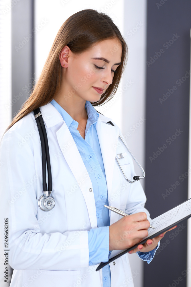 Doctor woman filling up medical form while standing near window in hospital office. Happy physician at work. Medicine and health care concept Doctor woman filling up medical form while standing near window in hospital office. Happy physician at work. Medicine and health care concept