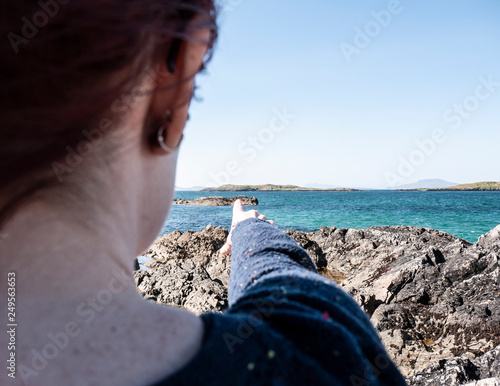 Over the shoulder shot of one woman pointing to a small island in the distance on a sunny day, with earrings and sunglasses. Taken on Renvyle beach along the Wild Atlantic Way in Ireland. photo