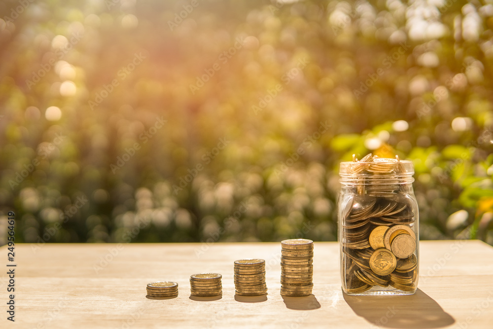 Coin and jar on wooden table in sunlight and blur background present the Growing Savings Coins - Investment And Interest and retirement Concept. Coin and jar on wooden table in sunlight and blur background present the Growing Savings Coins - Investment And Interest and retirement Concept.