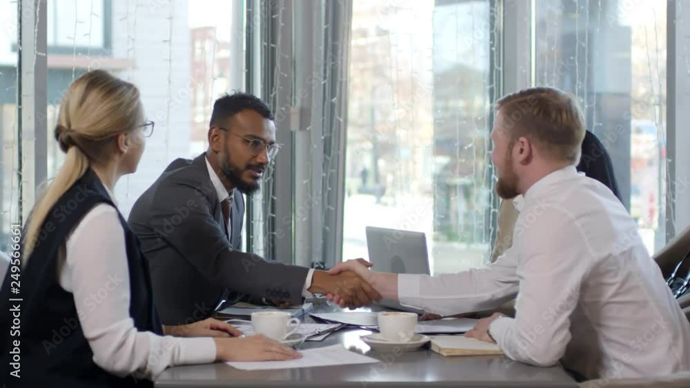 Multi-ethnic group of colleagues discussing contract at cafe table on break: middle eastern and Caucasian business partners smiling and shaking hands Multi-ethnic group of colleagues discussing contract at cafe table on break: middle eastern and Caucasian business partners smiling and shaking hands