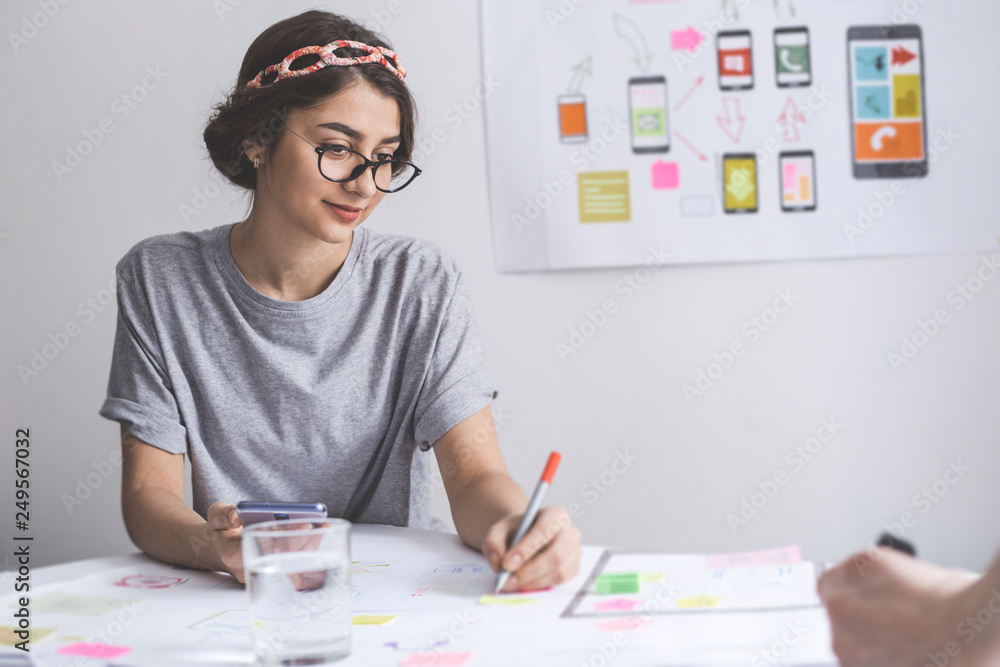 A girl designer of web applications for mobile applications, sits at the table and develops layouts and sketches with a partner. A girl designer of web applications for mobile applications, sits at the table and develops layouts and sketches with a partner.