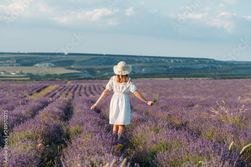 A girl in a white dress and a hat with bouquets in her hands stands back in a lavender field