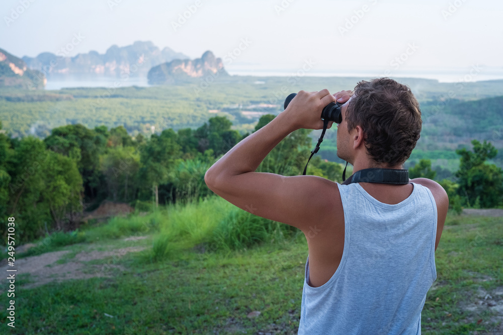A man looks through binoculars into the distance on the rocks and rainforest