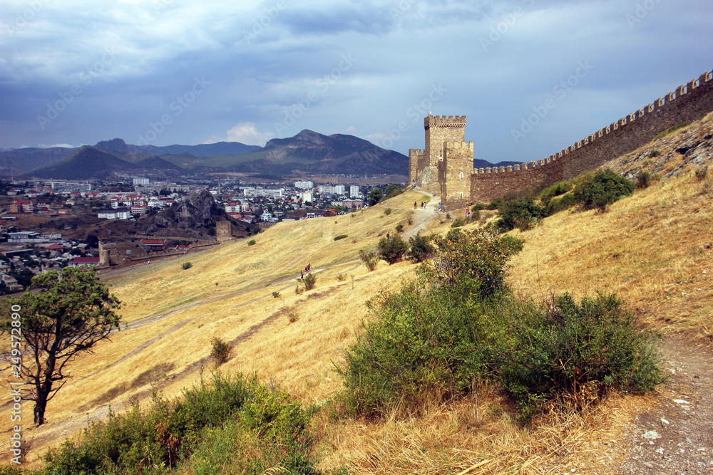 landscape with a stone fortress wall on a hill with yellow scorched grass on the background of mountains and sky