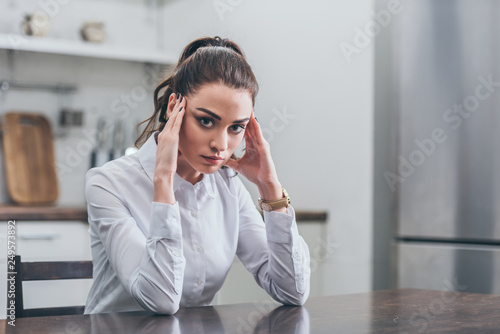 upset woman in white blouse sitting at table and putting hands forehead in kitchen, grieving disorder concept