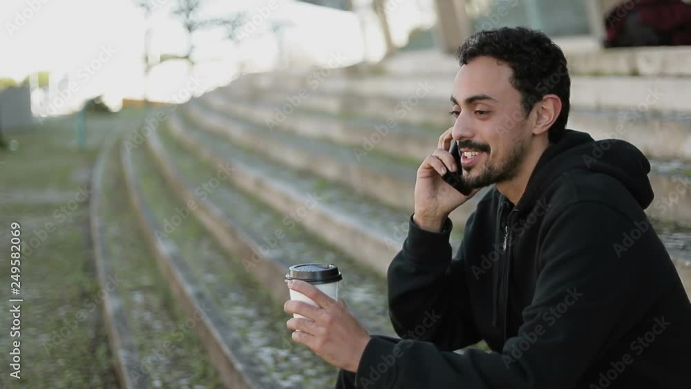 Side view of young Arabic man with dark curly hair and beard in black hoodie sitting on stairs outside, talking on phone, holding takeaway coffee in hand, drinking. Lifestyle, communication concept Side view of young Arabic man with dark curly hair and beard in black hoodie sitting on stairs outside, talking on phone, holding takeaway coffee in hand, drinking. Lifestyle, communication concept
