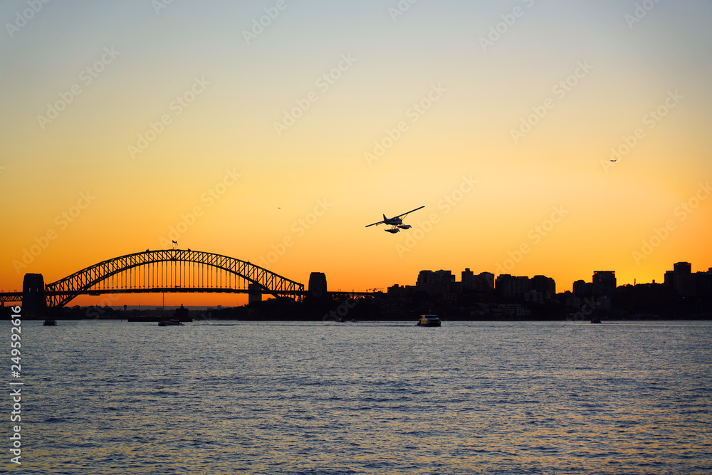 Naklejka premium Sunset view of a seaplane flying in the orange sky by the iconic steel Sydney Harbour Bridge in New South Wales, Australia