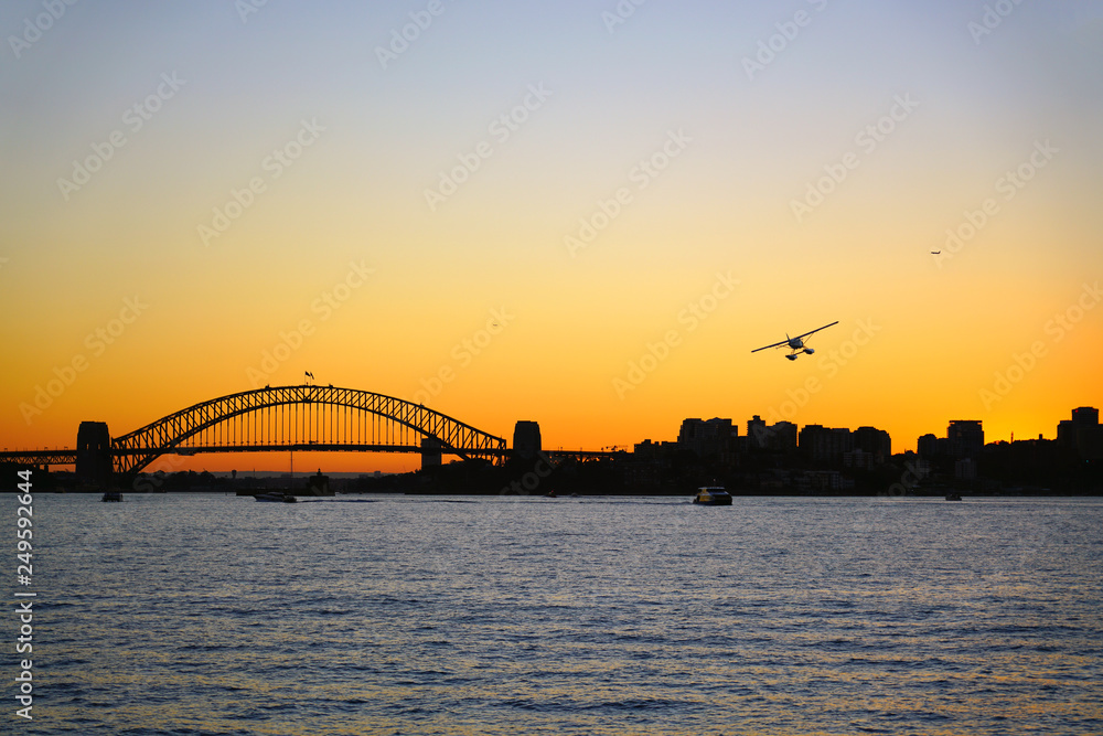 Naklejka premium Sunset view of a seaplane flying in the orange sky by the iconic steel Sydney Harbour Bridge in New South Wales, Australia