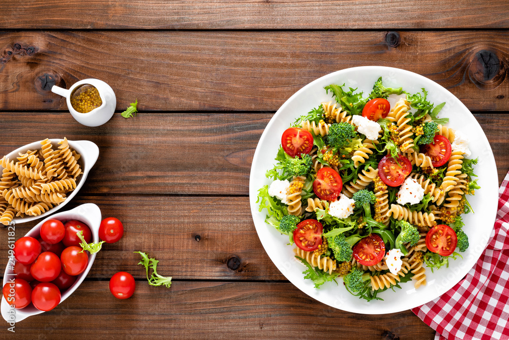 Italian pasta salad with wholegrain fusilli, fresh tomato, cheese, lettuce and broccoli on wooden rustic background. Mediterranean cuisine. Cooking lunch. Healthy diet food. Top view Italian pasta salad with wholegrain fusilli, fresh tomato, cheese, lettuce and broccoli on wooden rustic background. Mediterranean cuisine. Cooking lunch. Healthy diet food. Top view
