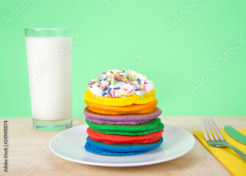 Stack of bright colorful pancakes topped with whipped cream and candy sprinkles  utensils and glass of milk . Green background. Fairy pancakes for National International Pancake day