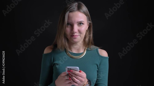 Portrait of young brunette caucasian girl positively nodding into camera and watching into smartphone on black background.