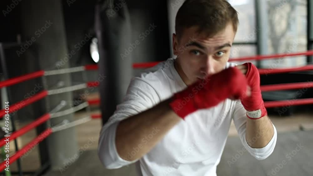 Confident boxer man wearing red bandages fights with shadow and doing some exercise at the boxing ring near the boxing pear in the loft place before the big fight. Confident boxer man wearing red bandages fights with shadow and doing some exercise at the boxing ring near the boxing pear in the loft place before the big fight.