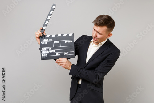Young business man in classic black suit looking on classic black film making clapperboard in hands isolated on grey wall background. Achievement career wealth business concept. Mock up copy space.