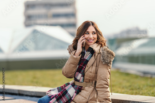 Beautiful Caucasian woman in jacket and with scarf talking on the phone while sitting on the bench outside on cold weather. © dusanpetkovic1